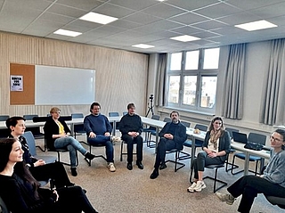Gruppe von Menschen sitzt im Stuhlkreis in einem hellen Raum mit großen Fenstern und einem Whiteboard an der Wand