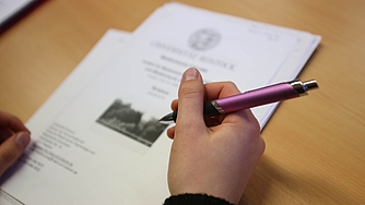 Hand hält einen Kugelschreiber in der Hand, auf einem Tisch liegt Papier